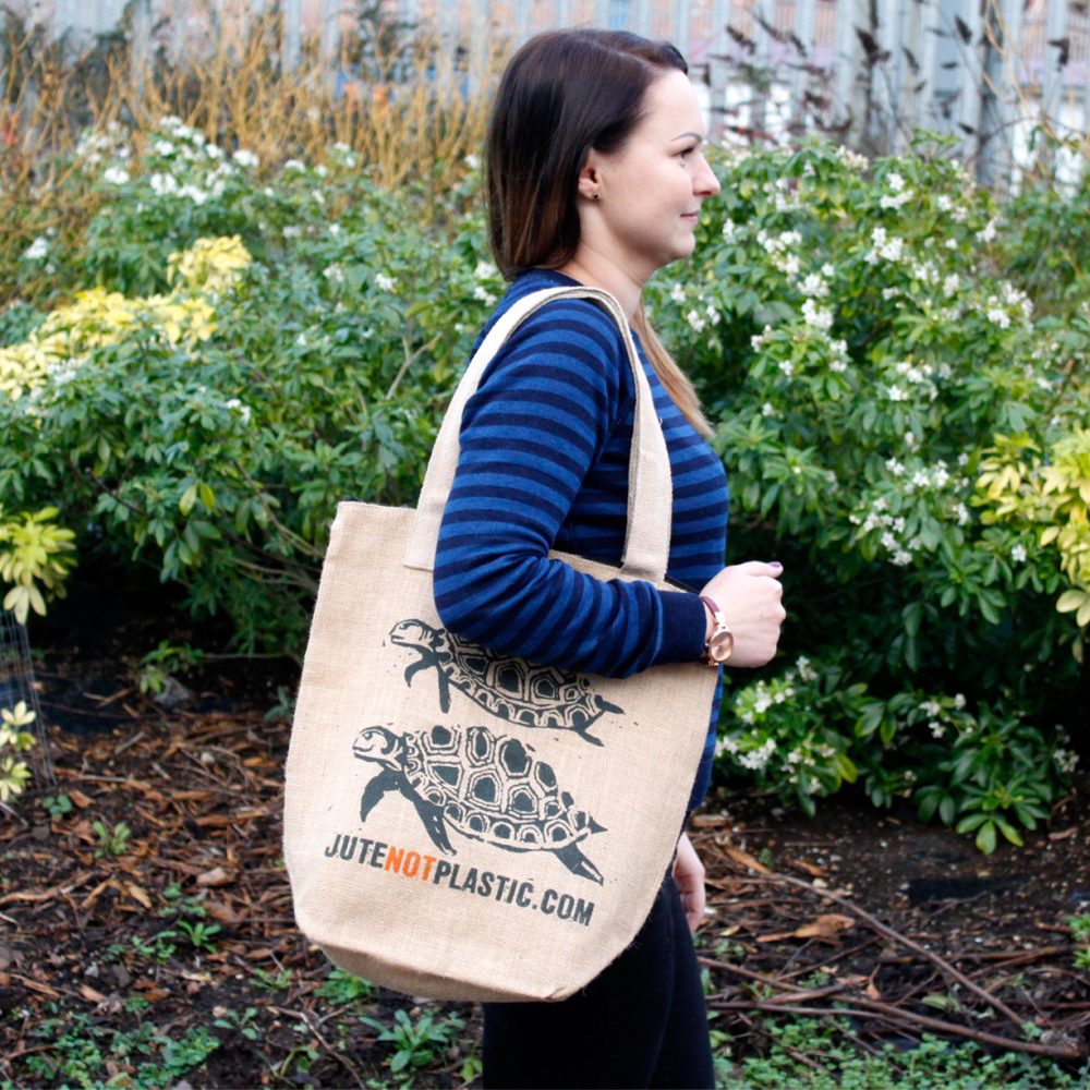 A woman carries a jute tote bag, printed with two turtles and the text "JUTENOTPLASTIC.COM," outdoors, in front of foliage.