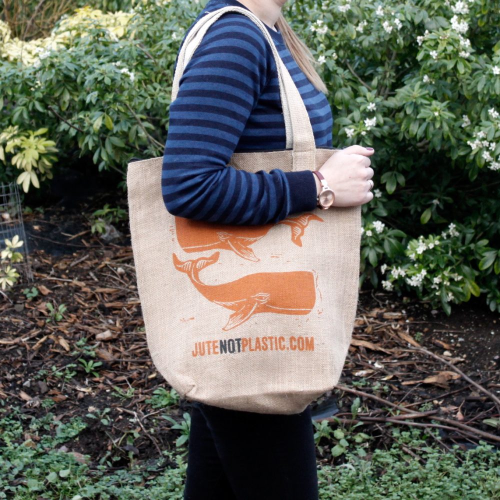 A person carries a tan tote bag with whale designs and "JUTENOTPLASTIC.COM" printed on it. They are standing outdoors near greenery.