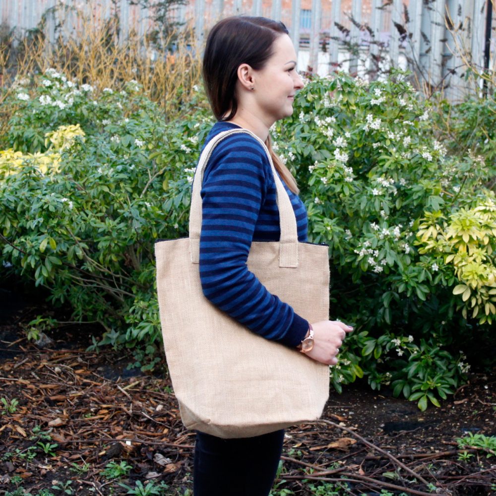 A woman carries a tan tote bag, outside by bushes and brown leaves on the ground.