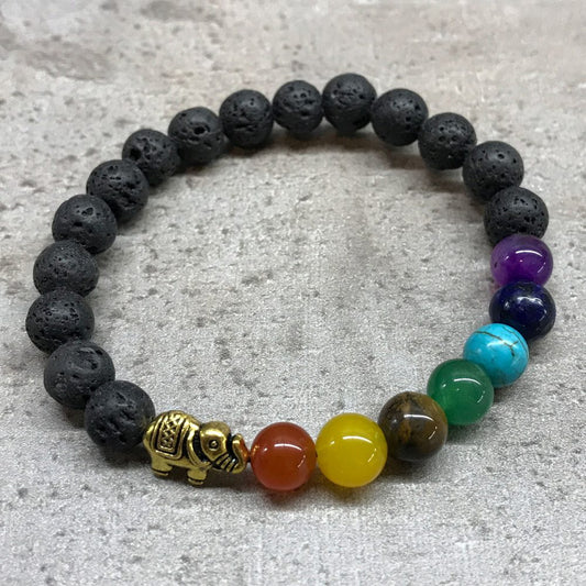 A stretch bracelet composed of black lava beads, seven chakra colored beads and a golden elephant charm, lays on a gray surface.