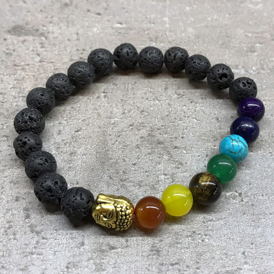 A beaded bracelet with porous black lava stones, chakra-colored stones, and a small golden Buddha head is presented on a gray surface.