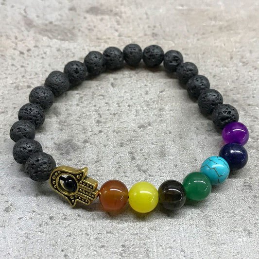 A beaded bracelet, mostly made of black lava beads and a hamsa hand charm, rests on a textured grey surface. Seven chakra-colored beads accent one part of the bracelet.