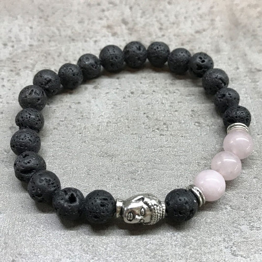 A beaded bracelet circles on a grey background. It alternates black porous lava beads and pink beads. It includes a silver Buddha head as an accent.