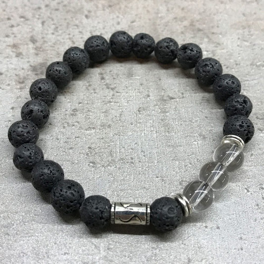 A beaded bracelet, made of porous black stones interrupted by silver metal and clear glass beads, lies in a circular shape on a gray surface.