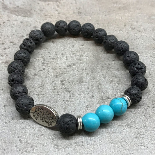 The beaded bracelet lies on a grey surface. It features porous black beads, three blue beads, silver spacers, and a silver charm with a leaf design.