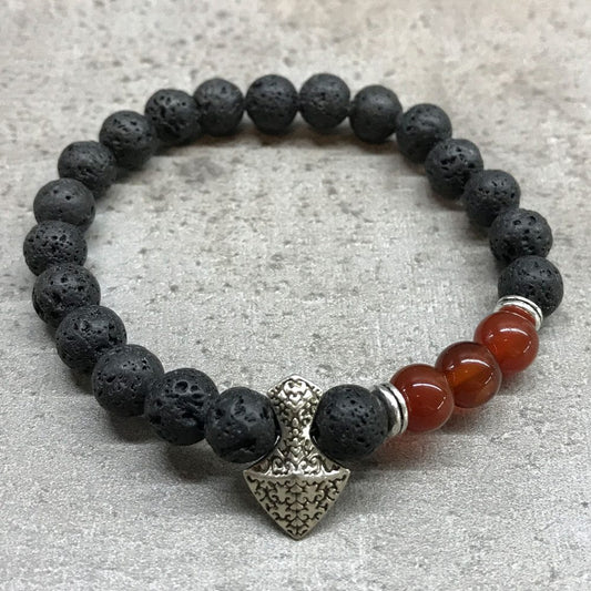 A beaded bracelet, made of black lava beads and carnelian beads and separated by silver beads, is on a textured gray surface. A silver charm in the shape of a hammer hangs from the beaded strand.