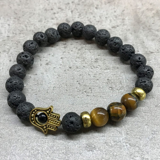 A beaded bracelet, composed mainly of black lava beads, is displayed on a gray textured surface, incorporating tiger-eye beads, gold-tone spacers, and a Hamsa hand charm.
