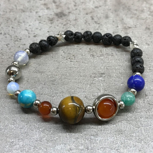 A bracelet of varied, colored stone and black lava beads, accented with small stars, rests on a grey, textured surface. The arrangement displays planets, represented by different colored stones.