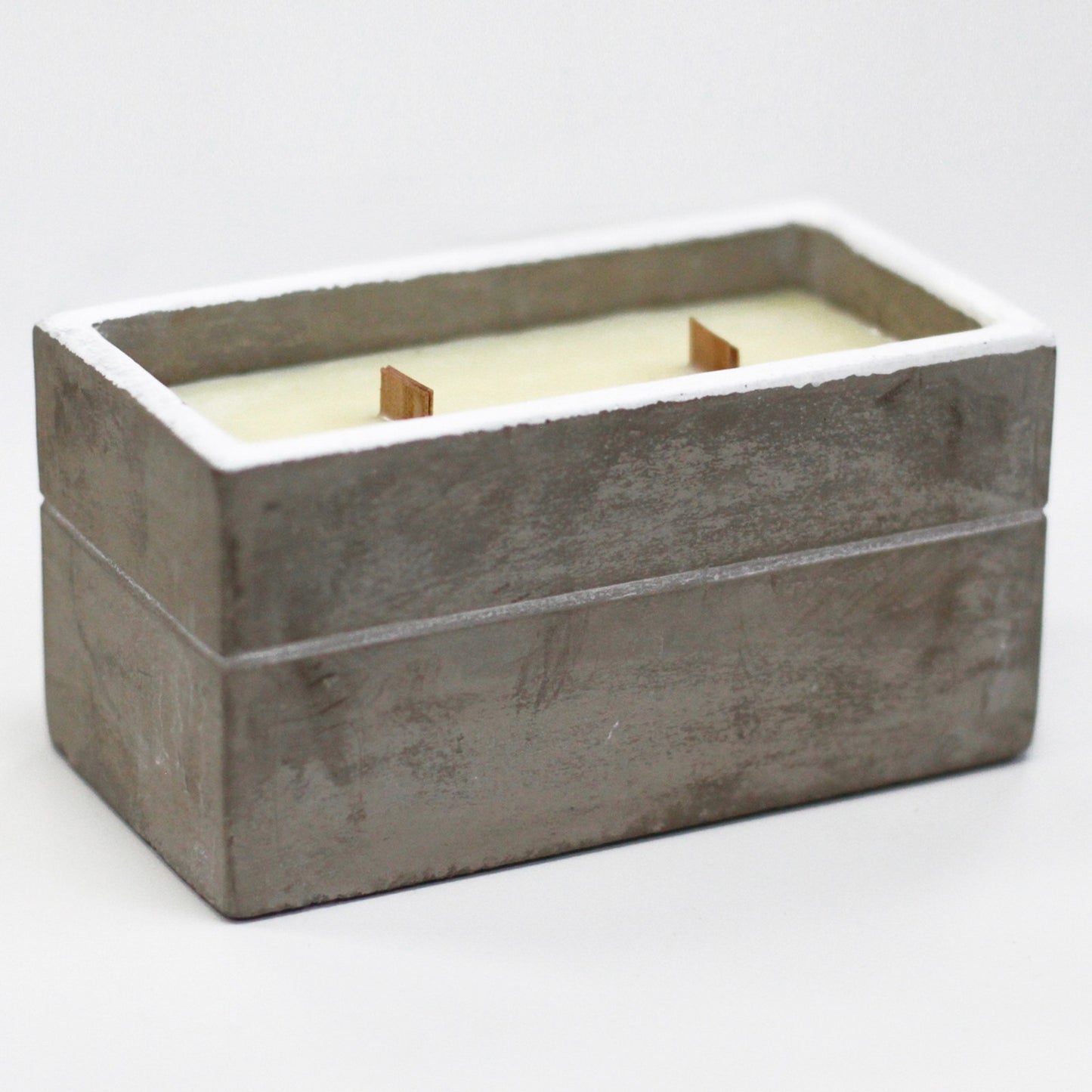 Concrete rectangular container holds pale wax with two upright wooden wicks, resting on a clean white surface against a soft out of focus light background