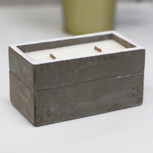 Rectangular concrete candle container holds smooth white wax with two short wooden wicks, resting on a white tabletop with a blurred yellow pot in the background.