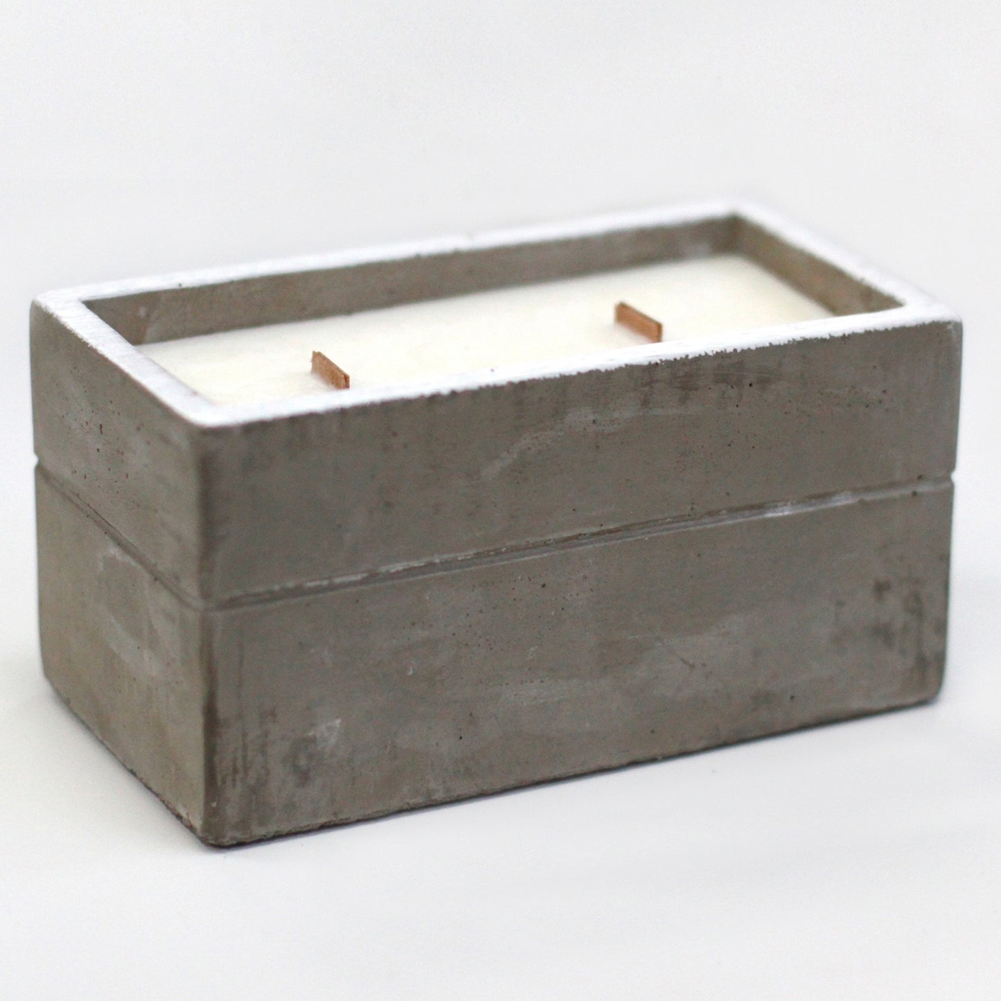 Rectangular concrete candle container holds white wax with two short wooden wicks standing upright on a plain white surface in soft studio lighting.