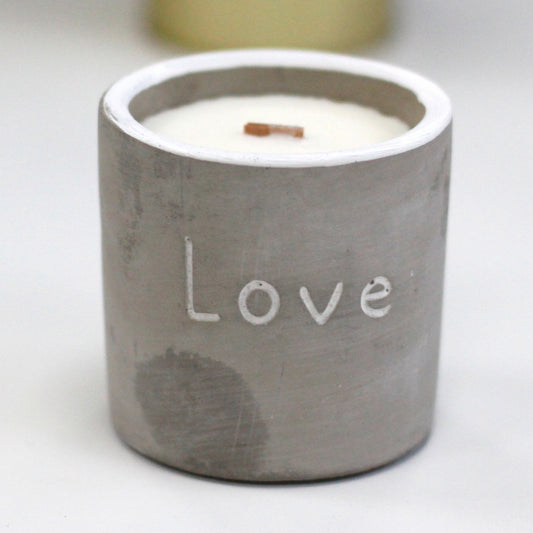 Concrete candle holder labeled Love sits on a white surface; unlit white wax with a small wooden wick visible, soft out-of-focus background.