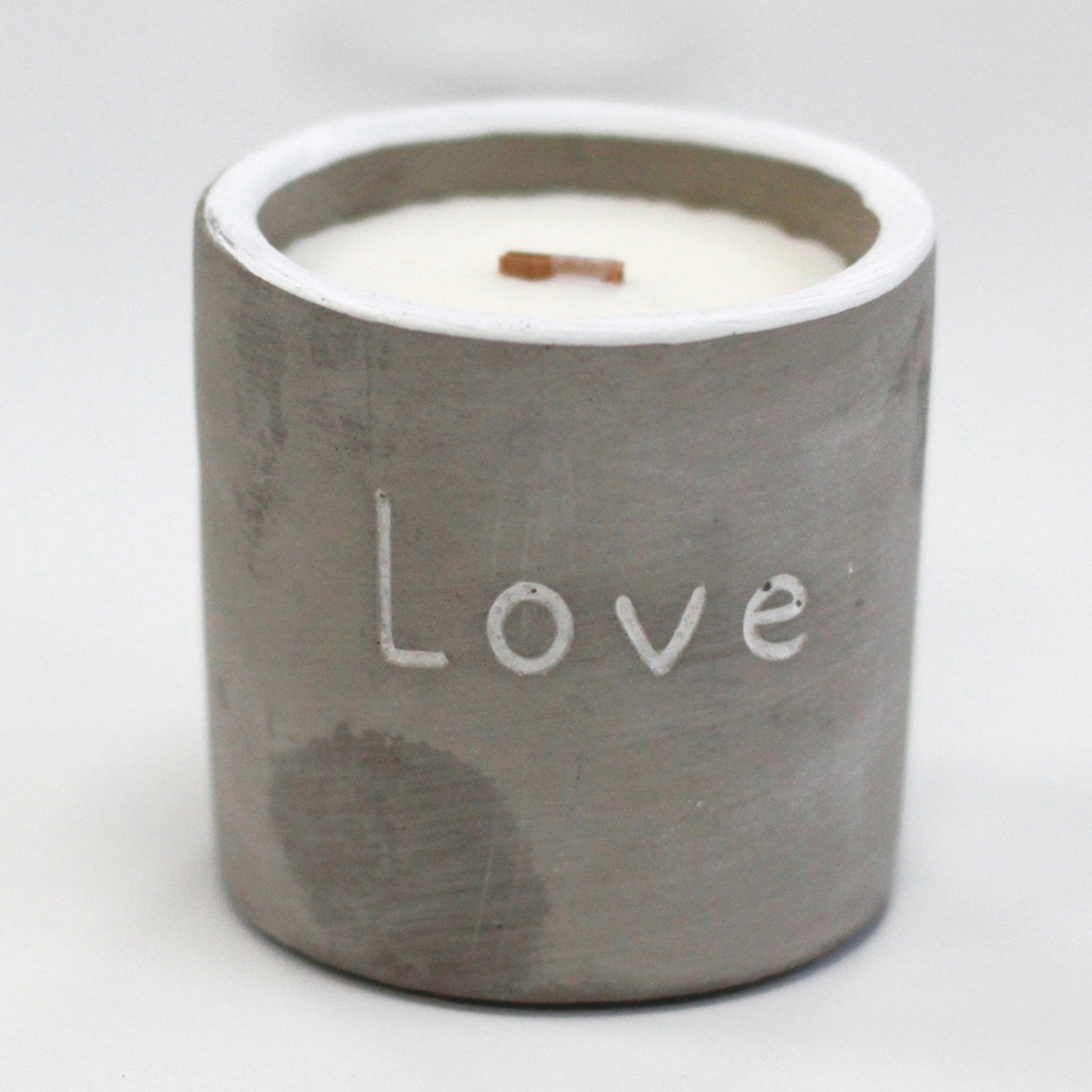 Small gray concrete candle container labeled "Love" holds white wax with a short wooden wick, standing on a plain white surface.