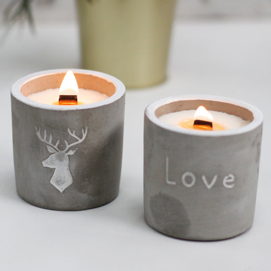 Two concrete candle holders burning on a white surface; left bears a deer silhouette, right reads "Love"; blurred plant pot in the background.