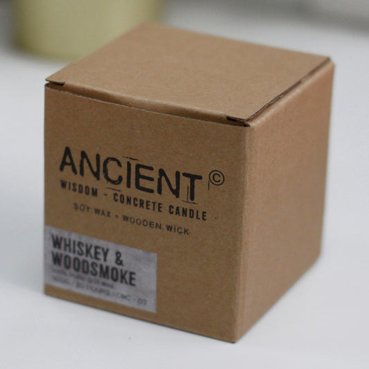 A small cardboard candle box sits on a white tabletop in soft light; front text reads ANCIENT © WISDOM - CONCRETE CANDLE SOY WAX • WOODEN WICK WHISKEY & WOODSMOKE