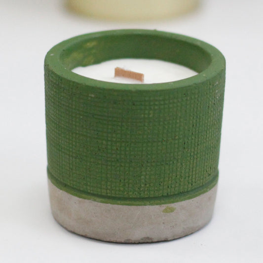 A small green textured cylindrical candle holder with a concrete base contains white wax with an upright wooden wick on a white surface with a blurred background.