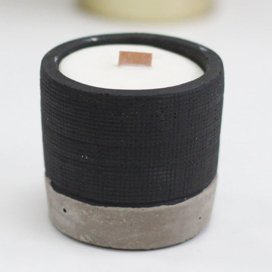A small cylindrical candle sits unlit with a short wooden wick in a textured black and raw concrete container on a white surface.