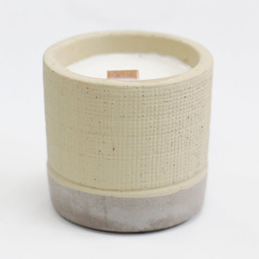 Beige textured ceramic candle pot holds an unlit white candle with a short wooden wick, the two-tone pottery vessel sitting on a plain white surface and background.