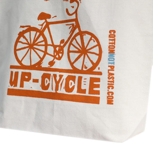 A white cloth bag shows an orange bicycle illustration above the word "UP-CYCLE", both printed in orange ink. "COTTONNOTPLASTIC.COM" is printed vertically on the side of the bag.