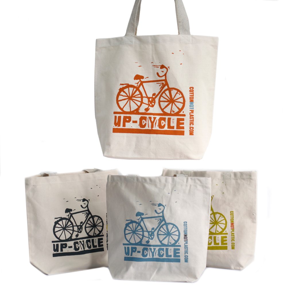 Four canvas tote bags feature bicycles with “UP-CYCLE” text printed beneath, and “COTTONNOTPLASTIC.COM” printed vertically on their right, with each in a different color.