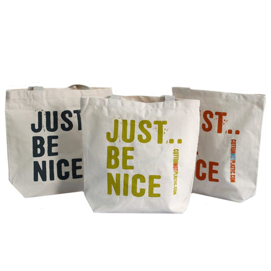 Three canvas tote bags stand in a row, each printed with the words "JUST BE NICE". On the right two, there's also “COTTONNOTPLASTIC.COM”. The text is printed in different colors.