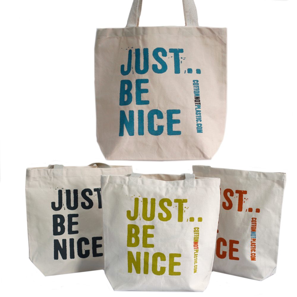 Four beige cotton tote bags stand in a group with the phrase "JUST... BE NICE COTTONNOTPLASTIC.COM" printed on them. The phrase is in blue, black, green, and orange on different bags.