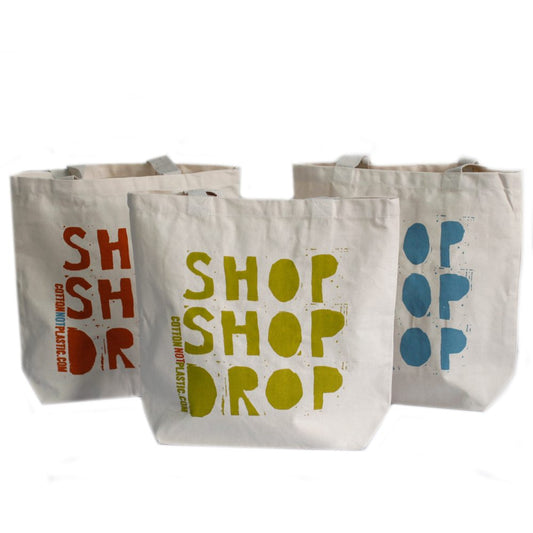 Three tan tote bags stand in a row against a white background. Each is printed with a stacked word design: “SH” in red with "COTTONNOTPLASTIC.COM", “SHOP SHOP DROP” in green, and “OP OP OP” in blue.