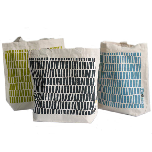 Three tote bags stand on a white surface. They are printed with rows of short rectangles in green, black, and blue, and bear the text "COTTONNOTPLASTIC.COM".