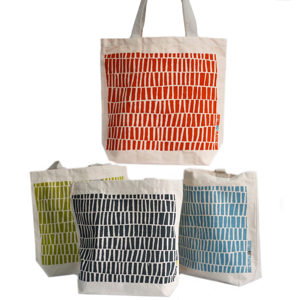Four canvas tote bags stand with different colors of repeating bars. The bags include red, green, black, and blue, and are labeled "COTTONNOTPLASTIC.COM". The background is white.