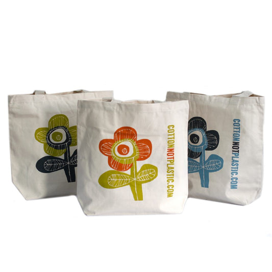 Three tote bags feature a flower design with the text "COTTONNOTPLASTIC.COM" vertically on the right. The first flower is olive/navy, the second is orange/olive, and the third is navy/blue, all on a white background.