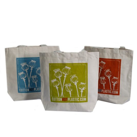 Three reusable cotton bags stand side-by-side; each displays poppy seed heads on a square colored background with the text "COTTONNOTPLASTIC.COM." The background colors include blue, green, and red.
