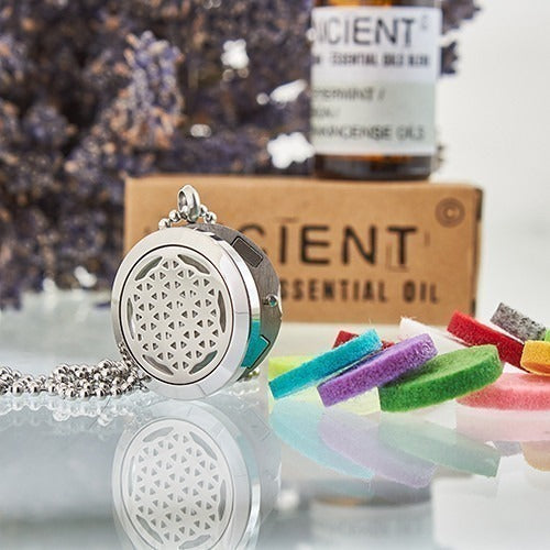 A silver pendant necklace rests on a reflective surface with colorful felt circles. Behind it are the texts: "ANCIENT ESSENTIAL BALM MIX Peppermint/ Frankincense Oils", and "ANCIENT ESSENTIAL OIL". Lavender and cardboard are in the background.