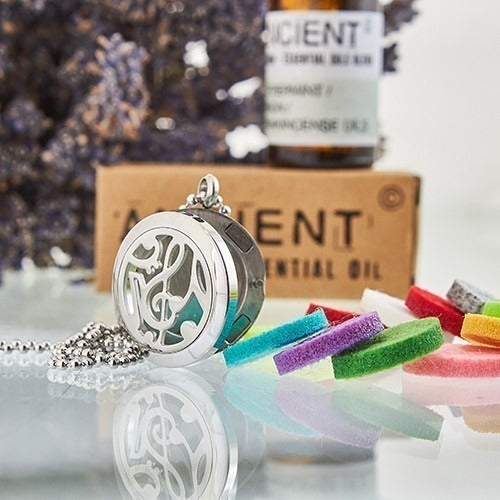 Aromatherapy necklace lies on a reflective surface, displayed with colorful felt pads and essential oil products. "ANCIENT ESSENTIAL OIL" is on a label. Dried lavender is in the background.