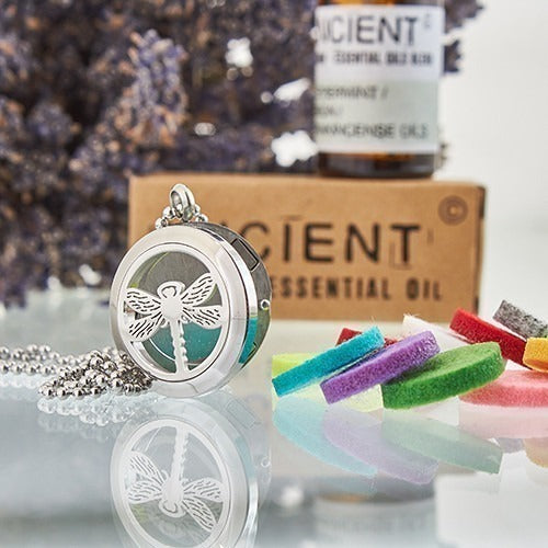 A diffuser necklace with a dragonfly design sits on a reflective surface with colorful felt pads; behind it, "ANCIENT" is printed on an essential oil box and bottle. Lavender buds are present in the background.