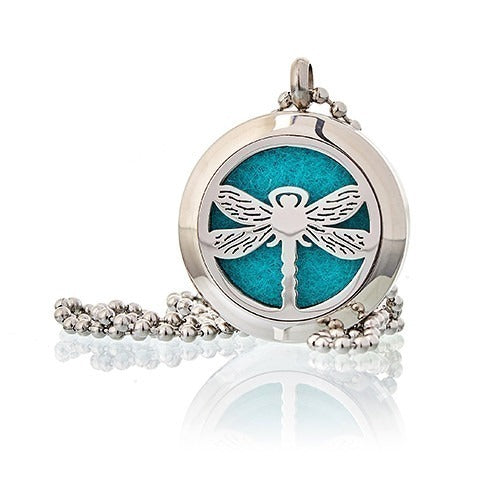 A silver, circular pendant with a cut-out dragonfly design displays a teal felt pad. It hangs from a beaded silver chain on a white reflective surface.