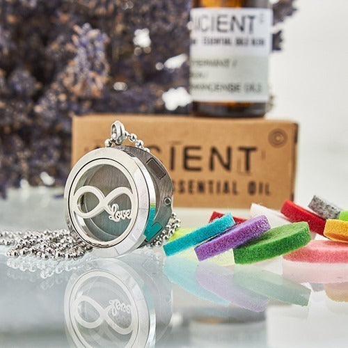 Silver pendant necklace sits on a reflective surface near colorful felt pads. Background items include a box that reads "IENT ESSENTIAL OIL" and a bottle with "CIENT" visible.
