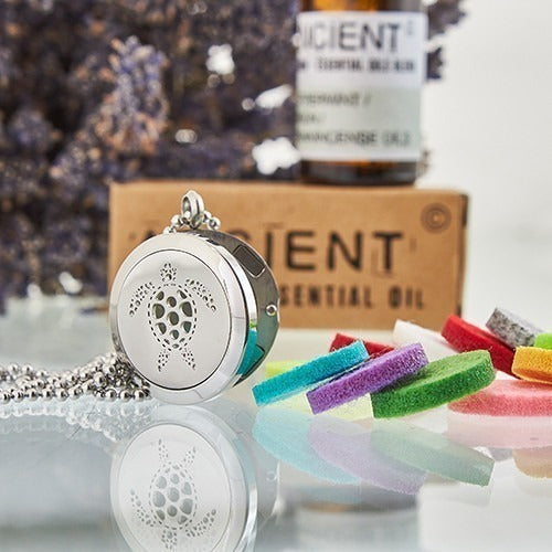 A silver turtle pendant sits with a chain, along with a scattering of colorful felt pads on a reflective surface. A brown bottle and cardboard box with "ANCIENT ESSENTIAL OIL" are behind the pendant.