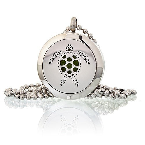 A silver, circular pendant with a turtle design sits on a beaded chain. The inside of the turtle shape is filled with green. The pendant reflects off a white surface.