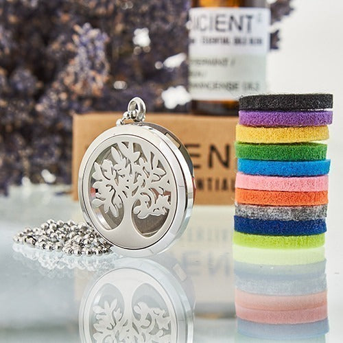 Silver tree-of-life pendant necklace sits on a reflective surface next to stacked, colorful felt discs. The background shows a bottle labeled "ANCIENT ESSENTIAL OILS".