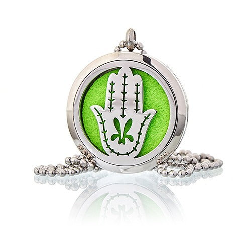A silver Hamsa pendant necklace reflects light on a white surface. The pendant's round case holds a bright green circle behind a white Hamsa design.