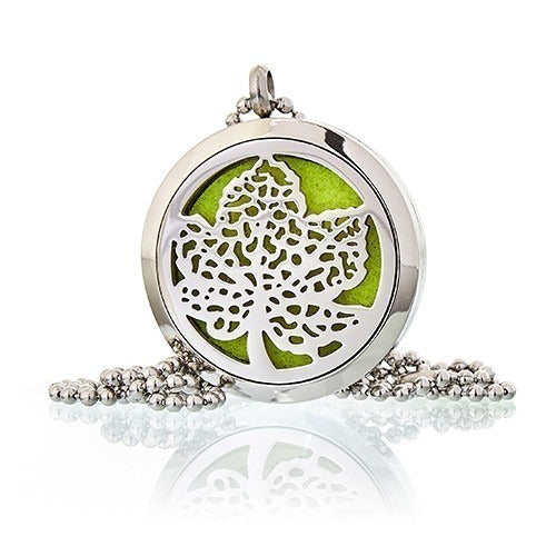 A round, silver pendant necklace features a cut-out tree design over a bright green background. The necklace rests on a white surface, with its reflection visible below.