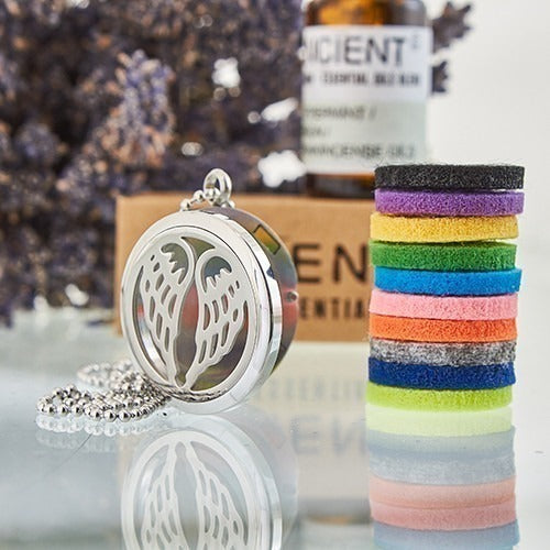 Aromatherapy necklace with wing cut-out lays on a reflective surface, with a stack of colorful diffuser pads, lavender sprigs, and a bottle labeled "ANCIENT" in the background.