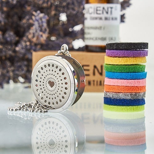 A silver, round, perforated locket necklace lies beside stacked, colored felt pads on a reflective surface. "ANCIENT" and "ESSENTIAL" are partially visible on objects in the background.