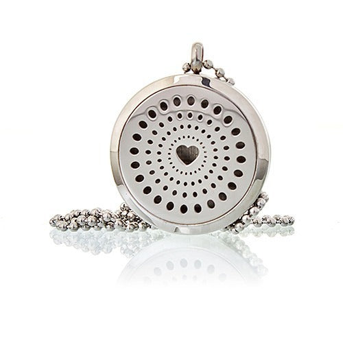 A silver locket with a circular design of small holes, arranged in a spiral towards a heart-shaped center, rests on a silver chain on a glossy white surface.
