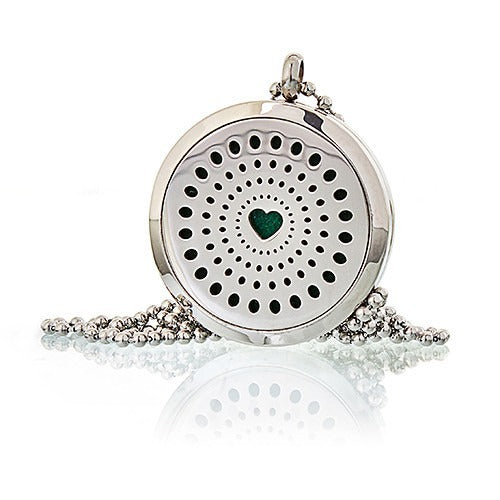 A silver pendant necklace lies still, displaying a circular filigree pattern surrounding a green heart, on a white surface that reflects its image.