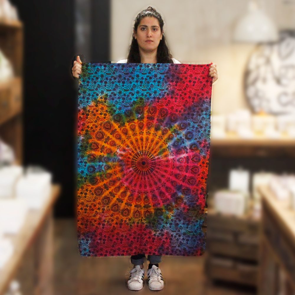 A woman holds a vibrantly colored mandala tapestry featuring swirling patterns in red, orange, blue, and teal. The tapestry is tie-dyed and has a detailed circular design. The context is a blurry indoor shop setting.