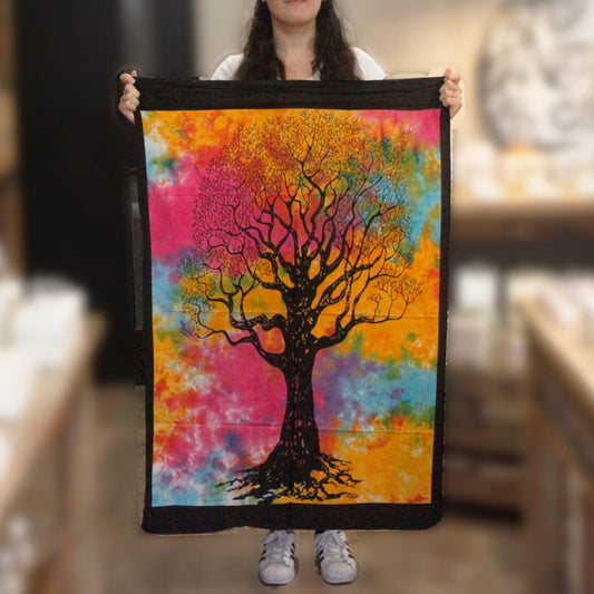 A tie-dye tapestry depicting a large tree is held by a person. The tapestry features a dark tree silhouette against a vibrant, multicolored background. The person is indoors.