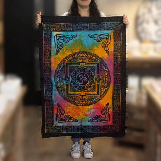 A tie-dye tapestry, featuring a mandala with the Om symbol, is held by a person. The tapestry is multicolored with a dark border. The setting appears to be a shop.