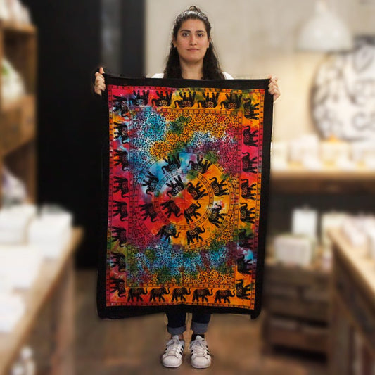 A woman holds a tie-dye tapestry featuring repeating black elephants. The tapestry displays a circular pattern in vibrant colors; it is being shown in a shop setting.