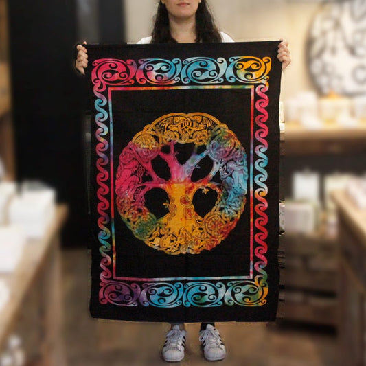 A person holds a tie-dye tapestry featuring a colorful Celtic tree of life encircled by knotwork. The tapestry is framed by a wavy border. The background is a blurred shop interior.